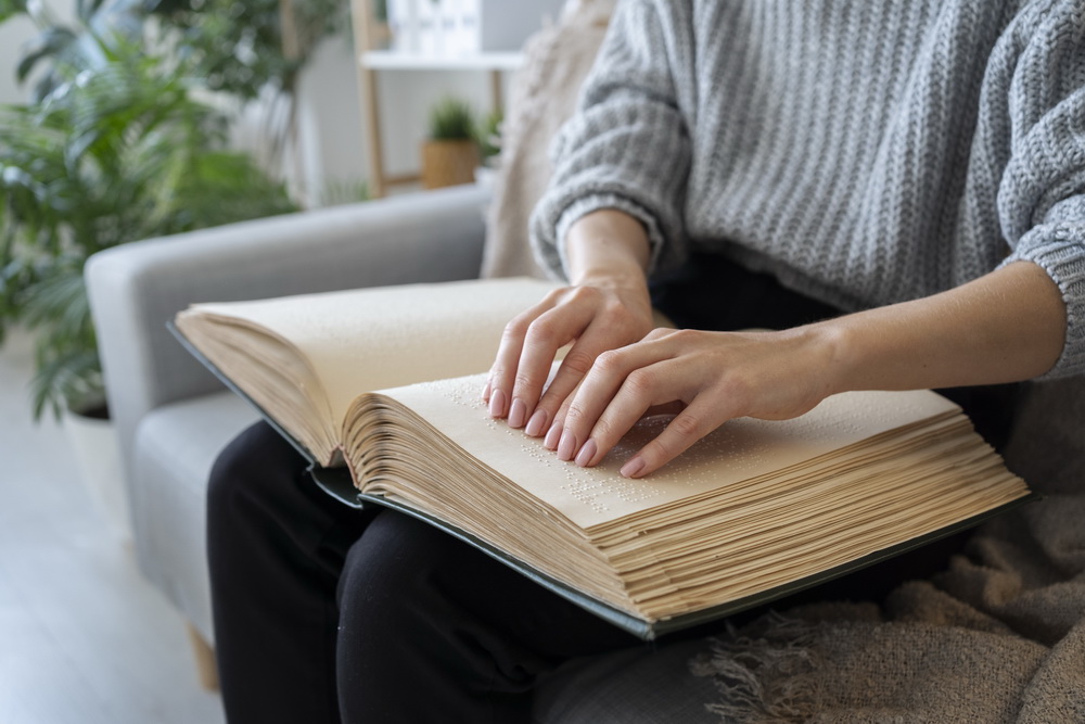 Auf dem Bild sieht man eine Person, die ein großes Buch in Brailleschrift liest. Die Person sitzt auf einem Sofa oder Sessel und trägt einen grauen, grob gestrickten Pullover und schwarze Hosen. Beide Hände sind auf den Seiten des Buches und tasten die erhabenen Braille-Punkte mit den Fingerspitzen ab. Die Seiten des Buches sind dick und cremefarben, was typisch für Braillebücher ist, da die erhabenen Punkte Platz benötigen.
Im Hintergrund sieht man eine gemütlich eingerichtete Wohnumgebung mit grünen Zimmerpflanzen, einem hellen Raum und Möbeln in sanften Farben. Die Szene vermittelt eine ruhige und konzentrierte Atmosphäre.. Auf dem Bild sieht man eine Person, die ein großes Buch in Brailleschrift liest. Die Person sitzt auf einem Sofa oder Sessel und trägt einen grauen, grob gestrickten Pullover und schwarze Hosen. Beide Hände sind auf den Seiten des Buches und tasten die erhabenen Braille-Punkte mit den Fingerspitzen ab. Die Seiten des Buches sind dick und cremefarben, was typisch für Braillebücher ist, da die erhabenen Punkte Platz benötigen.
Im Hintergrund sieht man eine gemütlich eingerichtete Wohnumgebung mit grünen Zimmerpflanzen, einem hellen Raum und Möbeln in sanften Farben. Die Szene vermittelt eine ruhige und konzentrierte Atmosphäre..